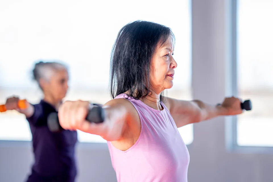 A focused middle-aged Asian woman in a pink tank top is doing arm stretching exercises with dumbbells in a bright studio. Meanwhile, other participants are working out in the background, highlighting the group fitness scene.