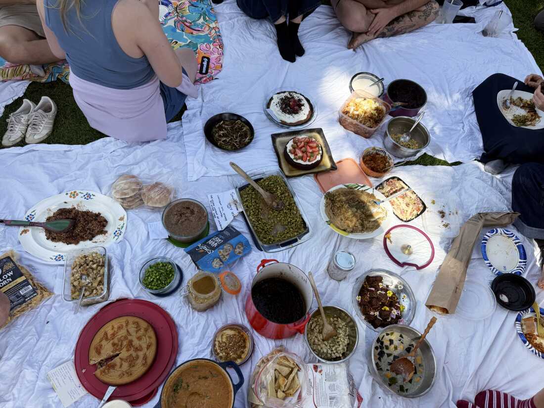 Several containers and pots containing plates of beans are placed on a white sheet spread on the ground. Several people are sitting on the grass near sheets and sheets.