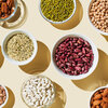 Various foods in bowls on a beige background. Foods include sources of dietary fiber such as mung beans, chickpeas, lentils, kidney beans, almonds, and hazelnuts.
