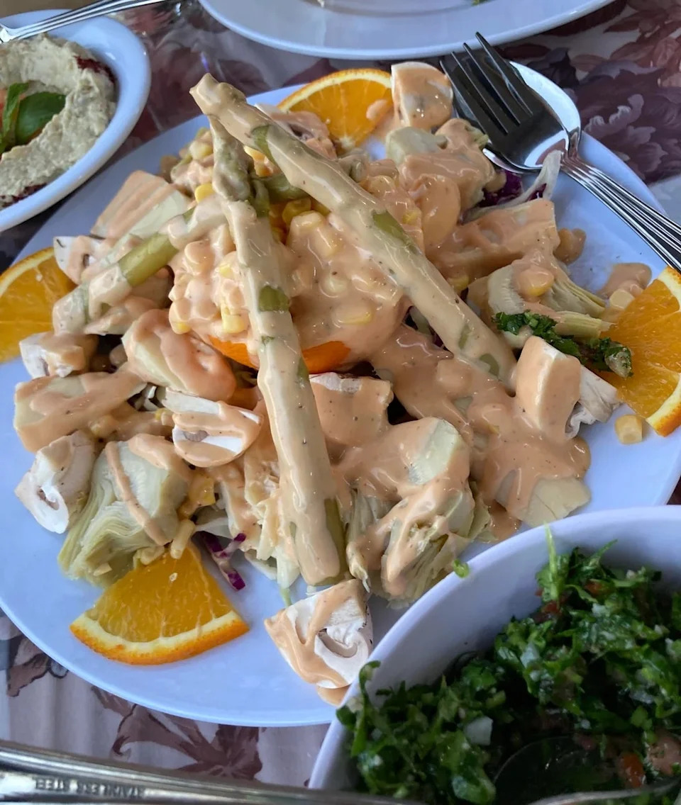 A plate of salad with asparagus, artichokes, orange slices and dressing. Served with hummus and tabbouleh in a small bowl