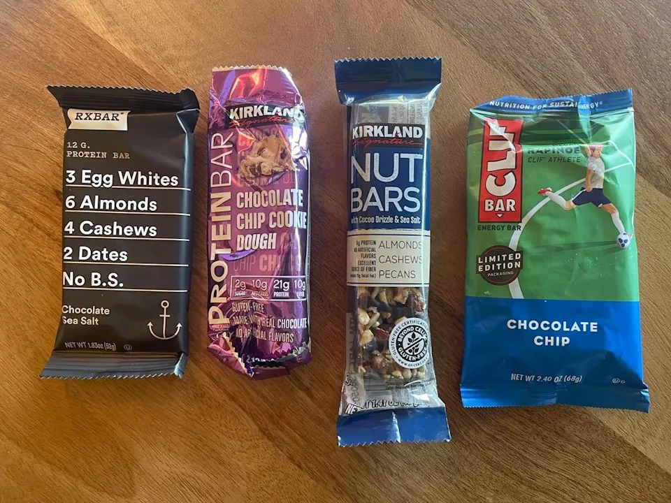 Four protein bars placed on a wooden surface: RXBAR, Kirkland Chocolate Chip Cookie Dough, Kirkland Nut Bar, and Clif Bar Chocolate Chip