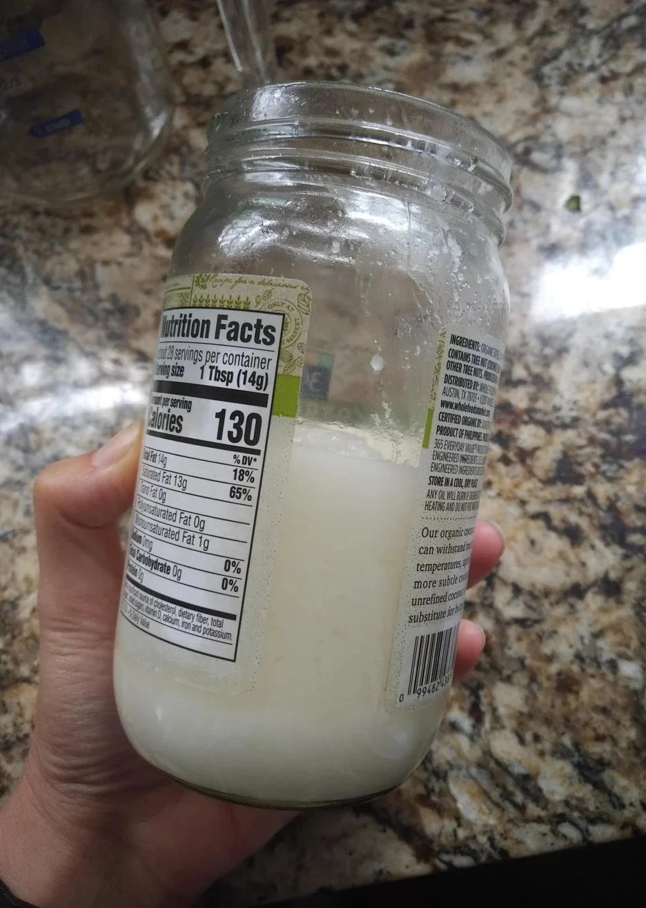 Hand holding jar of coconut butter with visible nutritional information label. The jar is placed on a marble countertop