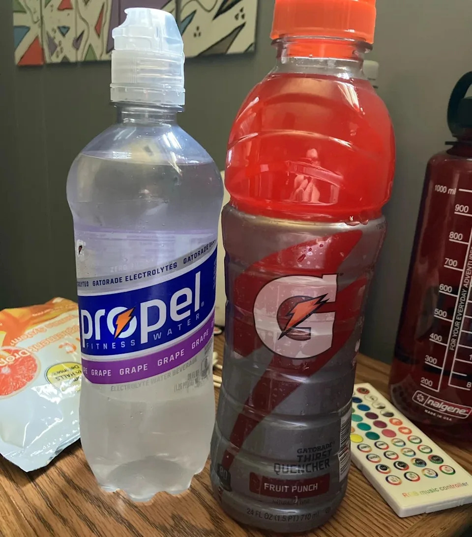 Two sports drinks on the table: a Propel Fitness water bottle and a Gatorade Fruit Punch bottle. Large water bottle and snacks in the background