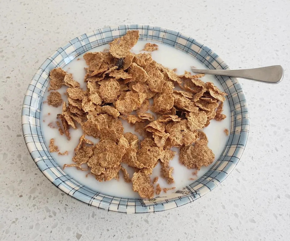 A bowl of bran flakes with milk on a checkered plate with a spoon in it