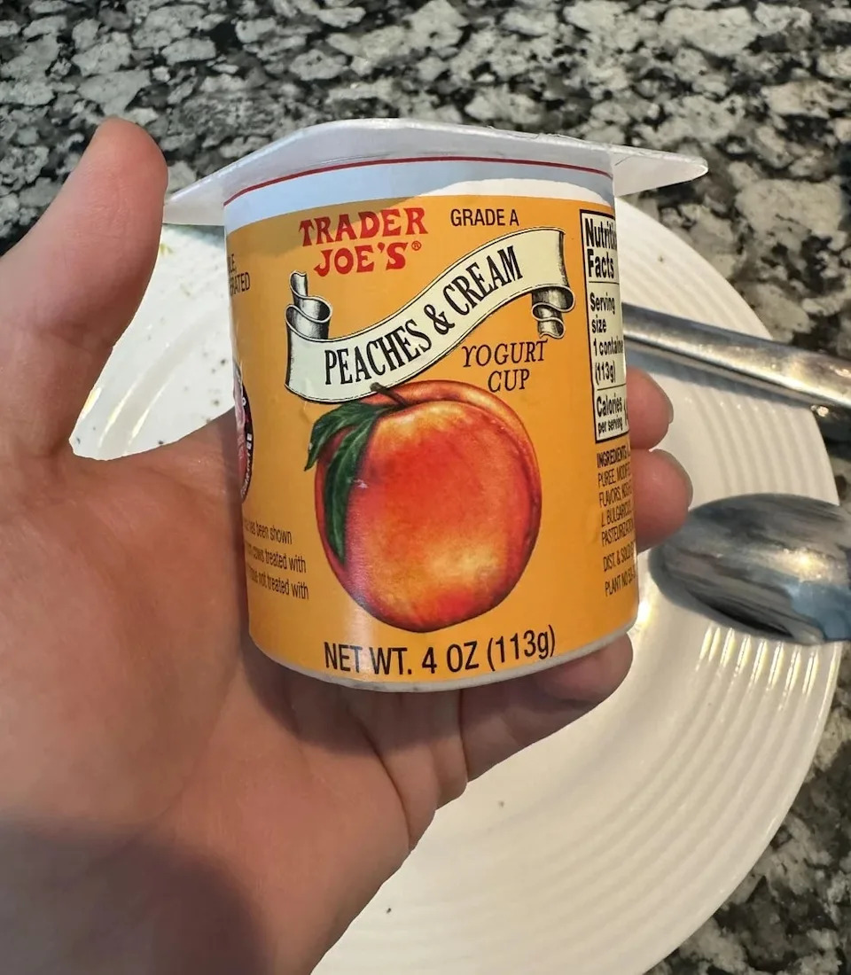 A person holds a Trader Joe's Peach and Cream yogurt cup on the kitchen counter with a spoon nearby.