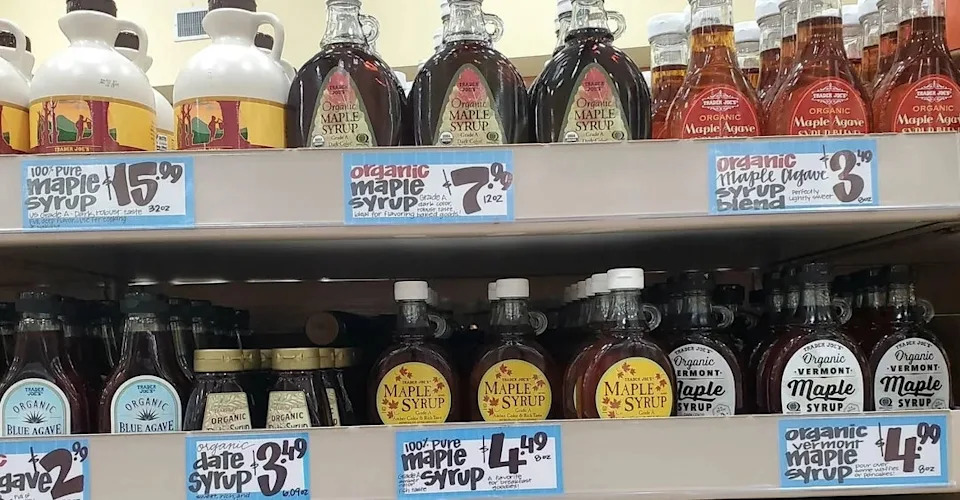 Various maple syrup bottles lined up on store shelves. Each has different prices and labels, including organic and Vermont options.
