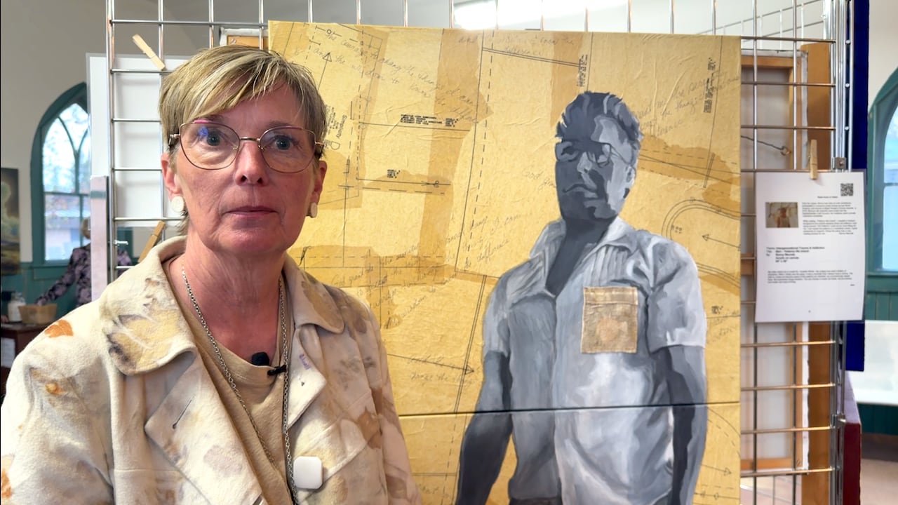 A blonde woman with short hair stands in front of a painting of a grayscale man against an old, yellowed sewing pattern.