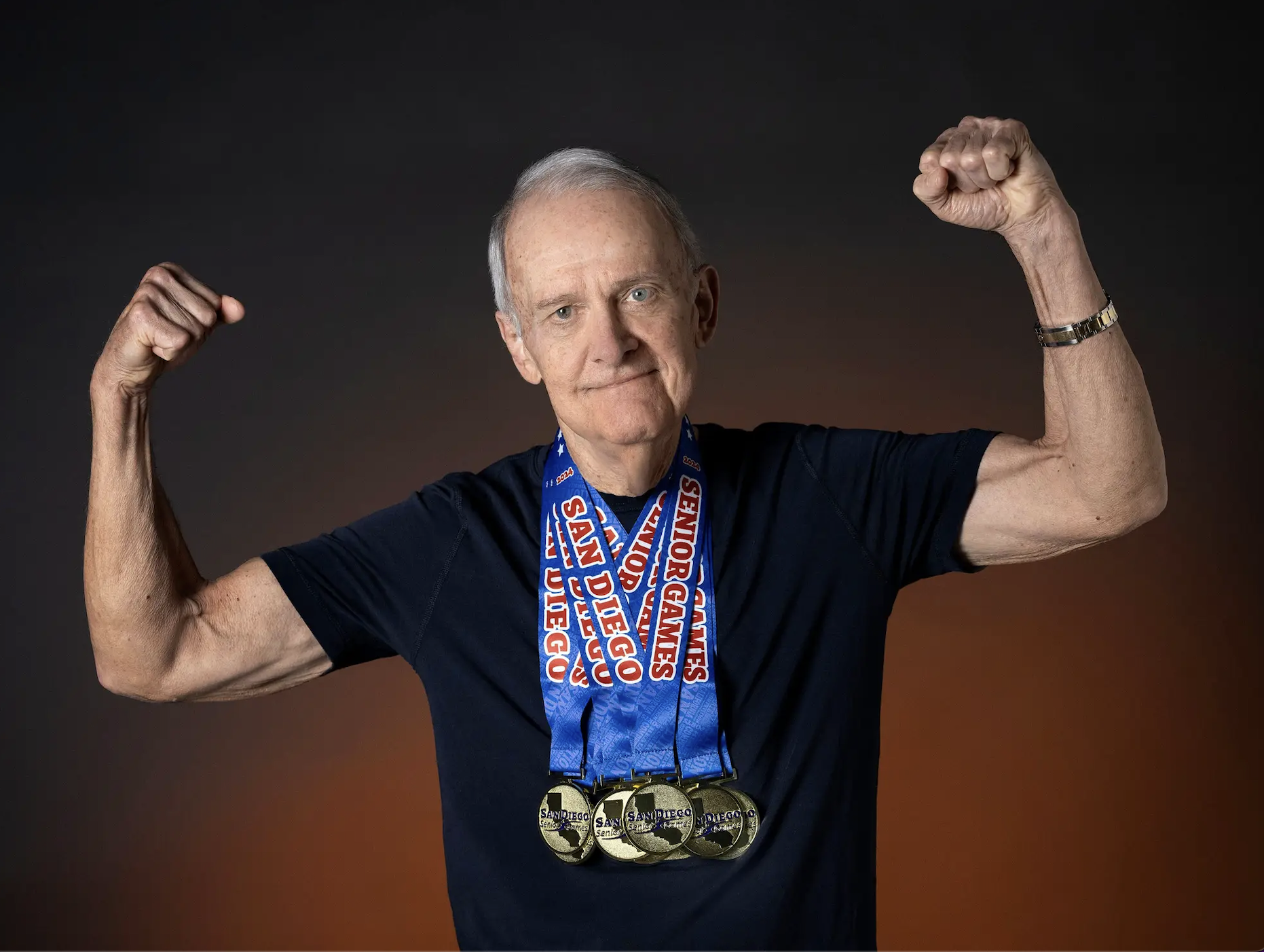 An older man posing flexing his biceps and wearing multiple gold medals "san diego senior games" It's written on the ribbon around her neck.