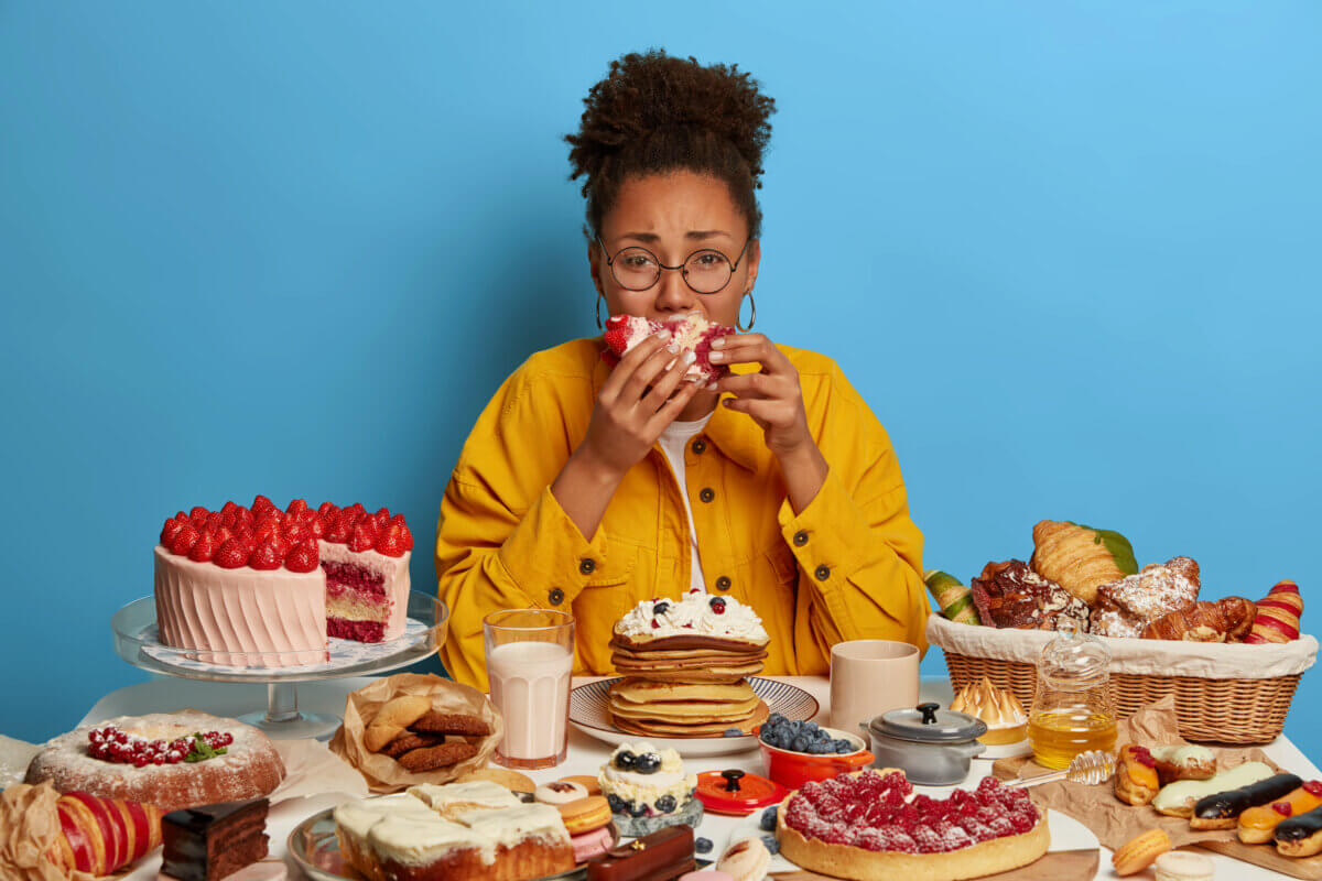 Gluttony and overeating concept. Upset and crying ethnic woman reluctantly eats cake, sits at table with many desserts, isolated over blue wall, feels hungry and greedy, wears yellow jacket