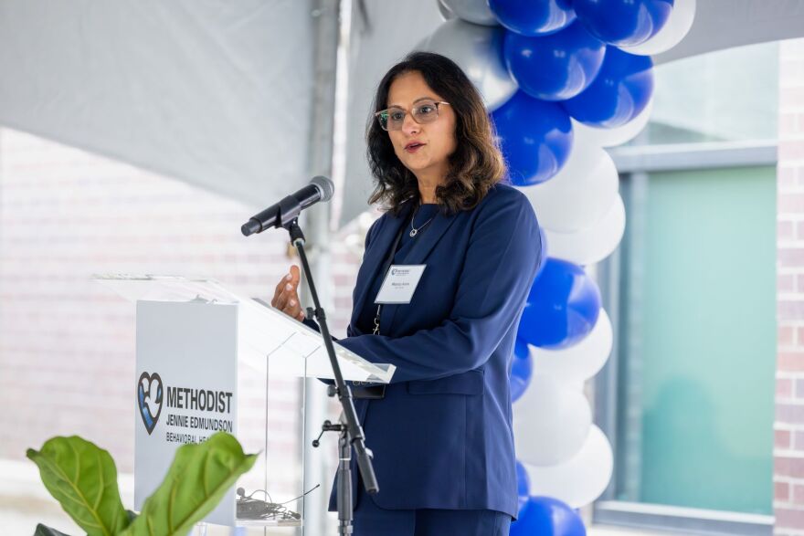 The photo shows the profile of a brunette woman with glasses wearing a navy blue suit. Others near her are out of focus.