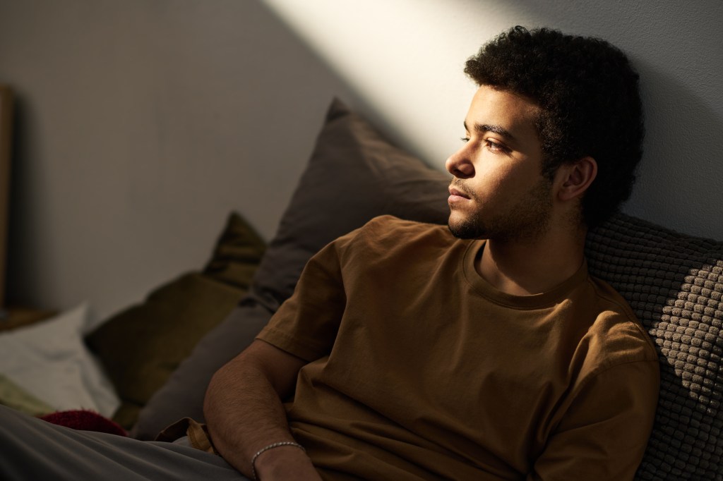 A sad teenage boy sits alone on his bed in his room, lost in thought.