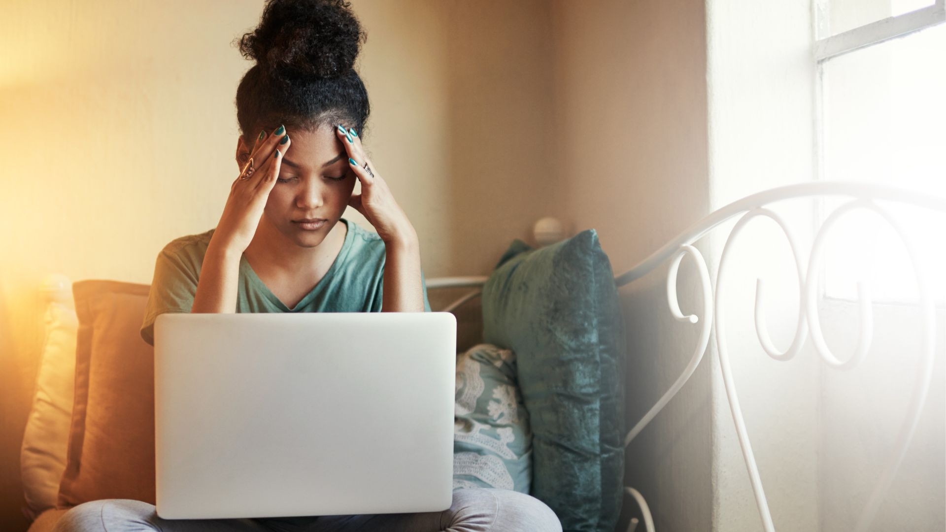 A young woman is sitting on the bed and putting her hands on her temples, looking stressed. She has a laptop on her lap.